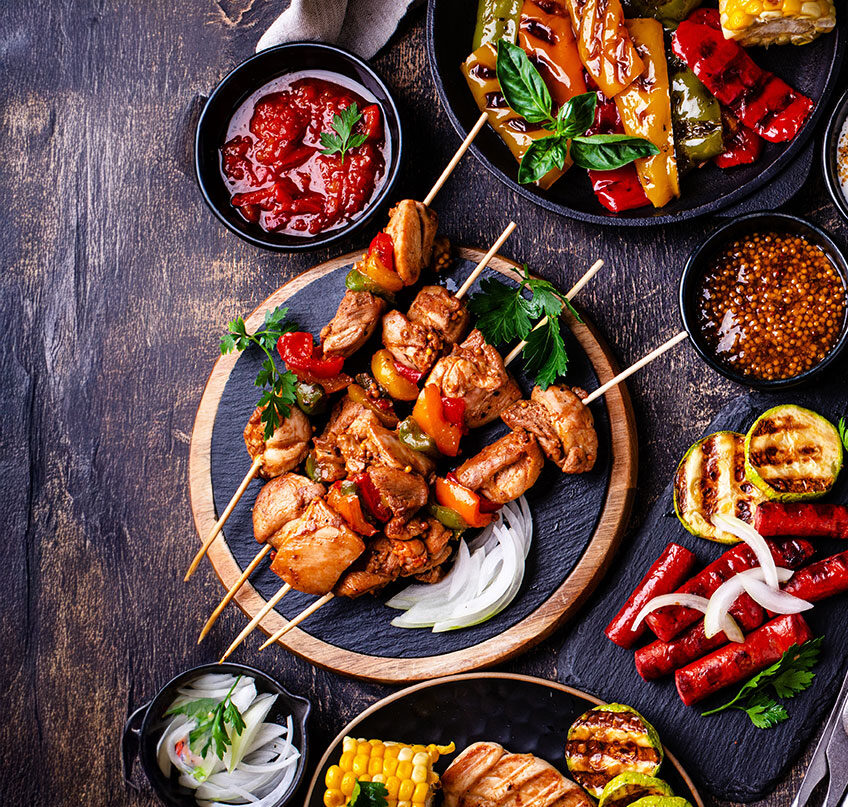 Grillteller mit Hähnchenspießen, gegrilltem Gemüse, Mais und verschiedenen Dips auf rustikalem Holztisch angerichtet.